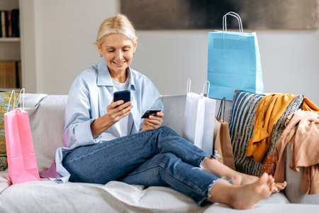 Joyful pretty caucasian mature blond stylish woman, happy with new purchased clothes, sitting on a sofa at home, using smartphone and credit card for online payments, many paper bags near, smilingの写真素材