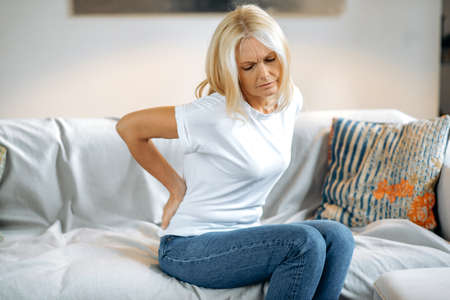 Upset frustrated mature woman suffering from backache, unhappy senior blonde sitting on a sofa at...