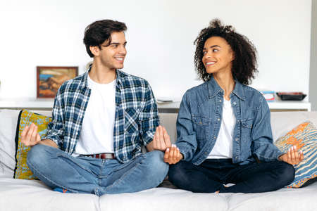 Multiracial happy couple on the couch at home. African American girl and hispanic guy, couple in love, in casual wear, sitting in lotus position on sofa, relaxing, meditate, look at each other, smileの写真素材