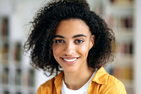 Close-up portrait of beautiful cute satisfied african american curly brunette girl, stands indoors,...