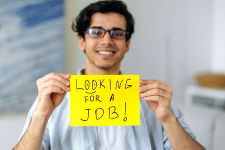 Defocused pleasant smart mixed race guy with glasses, student or unemployed, holding a sign with the inscription looking for a job, looking at the camera, smiling friendlyの写真素材