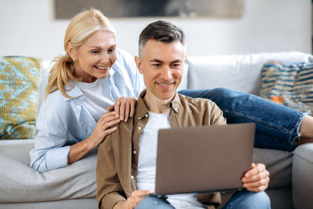Happy caucasian family couple, mature wife and husband at home on a sofa, in casual wear, using laptop, browsing internet, social networks, chatting with friends or family, looking for household goodsの写真素材