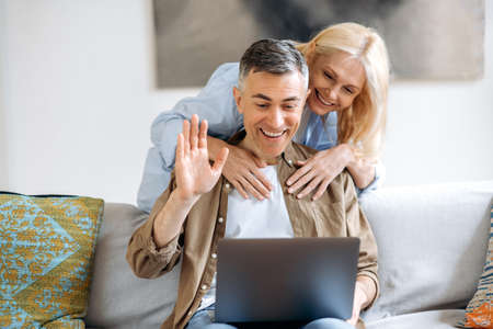 Video call communication. Happy senior caucasian married couple sitting on sofa in casual clothes, using laptop to chatting online with friends or family by video conference, greeting, smilingの写真素材