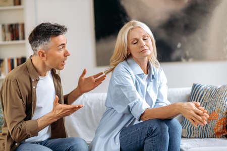 Misunderstanding, family quarrel. Upset, indignant gray-haired husband is emotionally talking and looking at his wife, gesturing with his hands, wife is upset because of a dialog, experiencing stressの写真素材