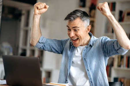 Happy successful lucky gray-haired caucasian man, freelancer or businessman, in casual clothes, works at home office, enjoying success, victory or good profit, looking at laptop, gesturing with handsの写真素材