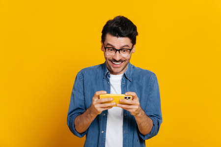 Photo of addicted caucasian male gamer with glasses, holds smartphone horizontally, plays online games, stylishly dressed, tries to win, spends free time, stands on isolated orange backgroundの写真素材