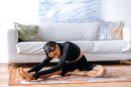 Flexible athletic young African American woman in sportswear, at home in the living room on the carpet, doing back stretching, doing yoga, leading a healthy lifestyle, closes eyes, smilingの写真素材