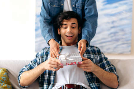 Surprised happy young Hispanic man sitting on the sofa in living room, in casual clothes, receiving...