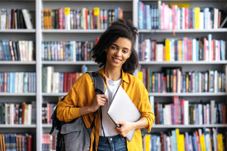 Portrait confident happy attractive teenage african american female student, standing against background of bookshelves in campus library, holding laptop and backpack, looks at camera, friendly smilesの写真素材