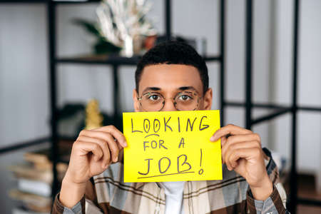 Unemployed smart mixed race hispanic guy with glasses, student or freelancer, holds a sign with the inscription looking for a job before his face, hopes to get a dream job, sadly looks at cameraの写真素材
