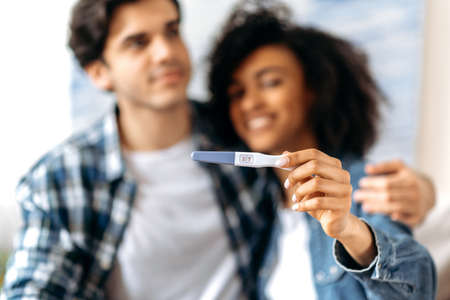 Joyful blurred young multiracial married couple in casual clothes enjoying the good news, getting ready to become parents, happy African American girl holds a positive pregnancy testの写真素材