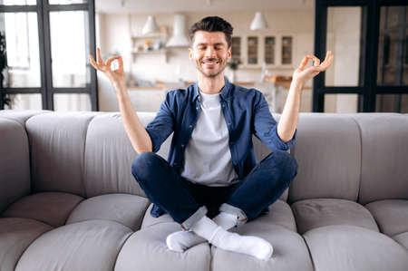 Meditation, relaxation, rest. Caucasian calm satisfied guy in stylish casual clothes sits on the sofa in the living room with closed eyes, meditates in the lotus position, relieves stressの写真素材