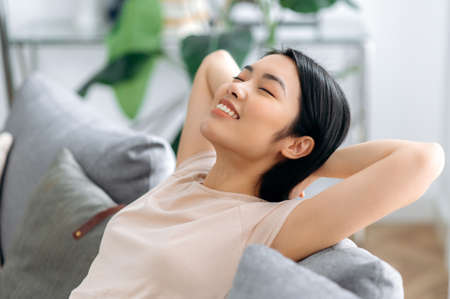 Side view of a satisfied young asian woman relaxes on a sofa in living room, smiling. Pretty...