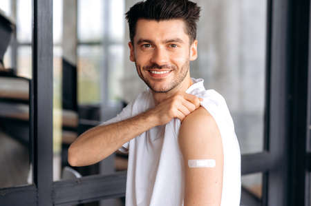 Vaccination and treatment during a virus or flu pandemic. Satisfied young caucasian man, wearing casual clothes, with adhesive plaster on shoulder after vaccination, looking at camera, smilingの写真素材