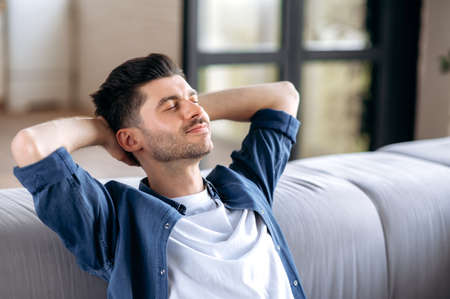 Close-up calm happy handsome caucasian bearded man in casual clothes relaxing on sofa at home, resting in living room, dreaming of vacation, put his hands behind his head, close his eyes, smilingの写真素材