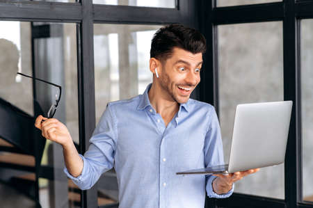 Surprised successful joyful caucasian business man or manager standing in the office, holding a laptop in his hand, communicating with colleagues via video conference, looking at the screen, smilingの写真素材