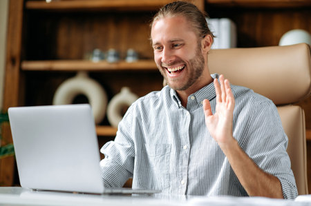 Confident friendly caucasian ceo, manager or business leader, sits at a work desk in the office, video chatting with a client or colleague using laptop, greeting with hand gesture, smiles. Video callの写真素材