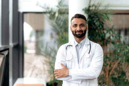 Portrait of an Indian general medicine doctor, stands with arms crossed in a clinic, dressed in medical clothes with a stethoscope around his neck, looking at camera, smiling friendly. Medical expertの写真素材