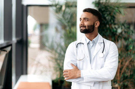 Indian handsome confident general medicine doctor, stands in a clinic, dressed in medical clothes with a stethoscope around his neck, looks away, smiling friendly. Doctor of the highest qualificationの写真素材