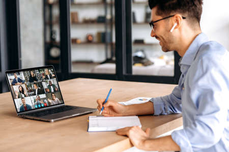 Side view to a confident male manager, taking notes during online business meeting with multinational colleagues, using laptop, sitting in office, planning marketing strategy, listening coworkersの写真素材