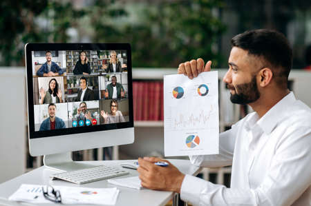 Video conference, online meeting. Intelligent Indian business man or mentor sits at workplace, communicates by video call with his partners, discuss a strategy, shows a financial graphの写真素材