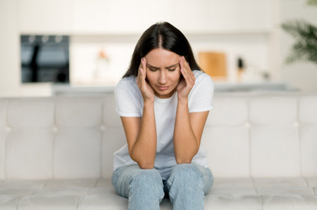 Sad caucasian brunette woman in stress, sitting on the sofa alone, in casual clothes, experiencing headache, migraine, massaging her temples, closing her eyes, need rest and treatmentの写真素材