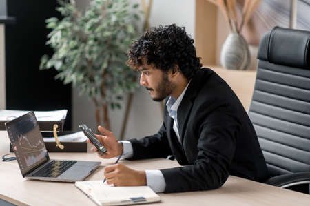 Side view of smart Indian crypto trader, investor, analyst broker, using laptop and smartphone...