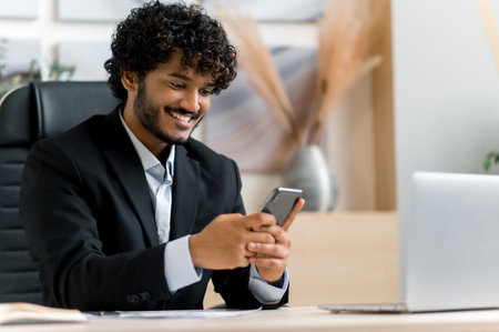 Online chat by smartphone. Satisfied Indian man, top manager, business owner sits at a desk, using a mobile phone while working, browsing Internet, answering email, texting with friends, smilesの写真素材