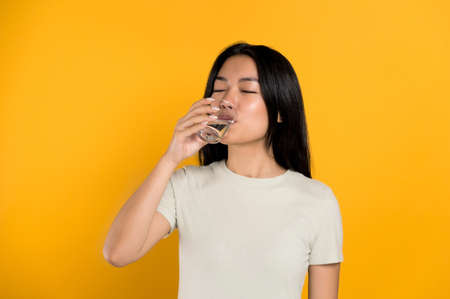 Healthy lifestyle concept. Positive, pretty chinese young woman in a t-shirt, drink a glass of clean water, cares of her health, drinks the daily norm of water, stands on isolated yellow backgroundの写真素材