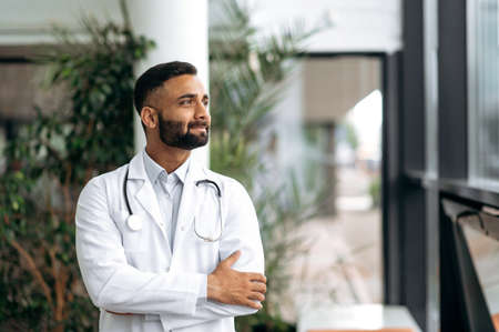 Portrait confident proud Indian professional therapist in medical uniform and stethoscope, standing in hospital on blurred background looking to side and smiling friendly. Medicine conceptの写真素材