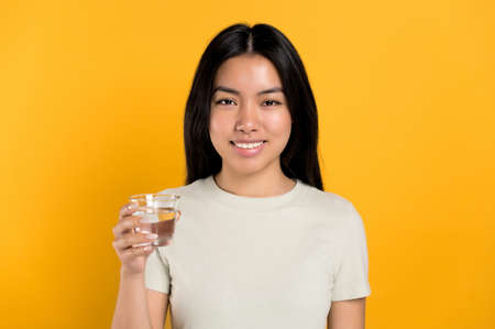 Positive, lovely chinese young woman in a t-shirt, holds a glass of clean water, cares of her...