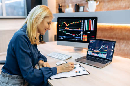 Successful smart mature woman investor and crypto trader, using pc and laptop, analyzes charts of trading in stock market and digital cryptocurrency exchange, conducts analysis, trading crypto coinsの写真素材