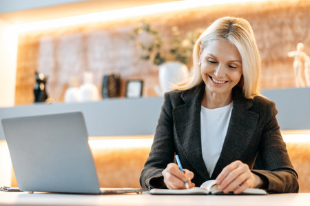 Happy smart caucasian businesswoman, tom manager, ceo, sitting at a table in a modern office, using a laptop, pondering business strategies and financial opportunities, taking notes, smilingの写真素材