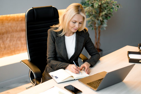 Successful smart influential mature focused confident caucasian blonde businesswoman, broker or financial expert, sitting at table in office with laptop, taking notes, develops business strategyの写真素材