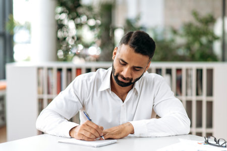 Concentrated serious smart confident Indian business man, manager or freelancer, wearing white shirt, sitting at table in modern office and taking notes, listening online webinar, brainstormの写真素材