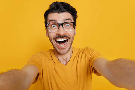 Funny caucasian guy in glasses, dressed in an orange T-shirt, makes a selfie, fooling around at the camera, smiling, standing on an orange backgroundの写真素材