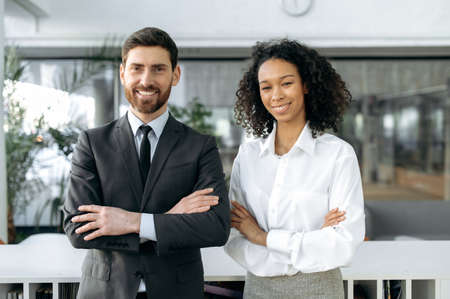 Influential successful coworkers of different nationalities, caucasian man and african american woman, in formal clothes, are standing with arms crossed in modern office, looking at camera and smilingの写真素材