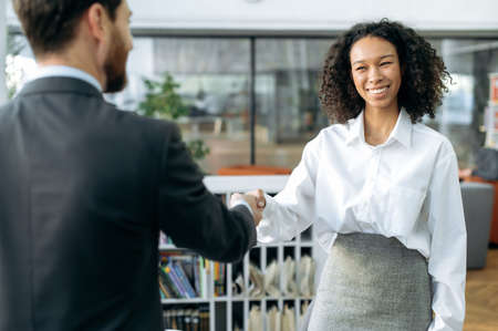 Making a deal, shaking hands, confirms agreements. African american successful business woman, sales manager, shakes hands with male colleague in modern office, smiling at each otherの写真素材