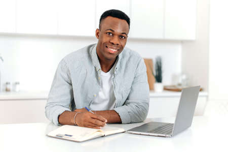 Positive modern black man, freelancer, designer or student, sits at a kitchen at home, using laptop, studying or working online, improves qualifications, smil friendly, takes notes. E-learning conceptの写真素材