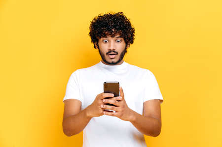 Surprised shocked Indian guy in white t-shirt, uses his smartphone, looks in amazement at screen,...