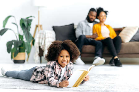 Happy African American family. Little cute African American girl, preschooler, lies on the floor in living room with a book in hands, looks at camera, smile, parents on the background sit on the couchの写真素材