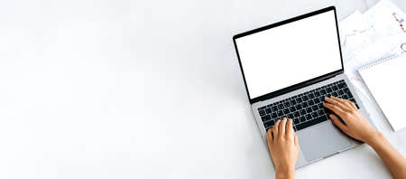 A laptop with a blank white screen, financial charts and a notepad lie nearby. Female hands on the...