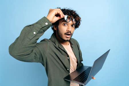 Shocked amazed Indian or Arabian guy, in casual wear, take off glasses, holds an open laptop in hand, looks surprised and confused at the camera, standing on isolated blue backgroundの写真素材