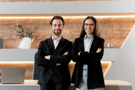 Successful intelligent caucasian young adult man and woman, office colleagues, financials, entrepreneurs, standing at the desktop, in formal and stylish clothes, arms crossed, friendly smilingの写真素材