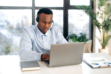 Smart purposeful, focused African American young man in headphones, manager, support worker, online consultant, sitting at a laptop in the office, having an online consultation with clients in a chatの写真素材