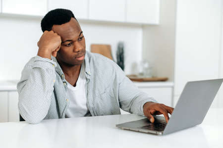 Bored frustrated african american guy, freelancer, designer, sitting at a table at home, dressed in casual clothes, working at a laptop, tired, in needs of rest or break, upsetの写真素材