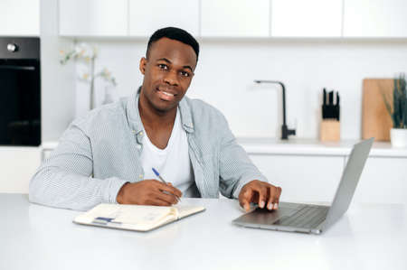 Attractive African American guy, student, freelancer, sits at a table in the kitchen, uses laptop and notepad for online learning, looks at the camera, listens to a lecture, takes notes,smile friendlyの写真素材