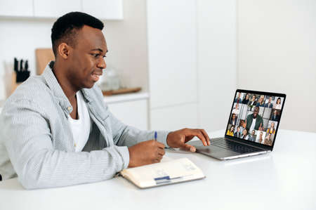 Smart positive african american man sits at a table at home, using a laptop, takes part in a multi-rice video conference with colleagues or employees, takes notes. E-learning concept. Work from homeの写真素材