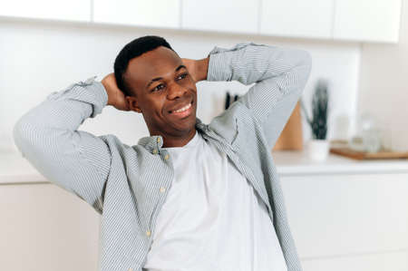 Portrait of a handsome African American man. Black attractive guy dressed in stylish casual clothes sitting at home in the living room in a relaxed pose with his hands behind his head, smiling happilyの写真素材