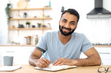 A smart positive Indian man, sitting at a table in the kitchen at home, listening to a lecture, webinar via video conference, taking notes in a notebook. Distance learning, advanced trainingの写真素材